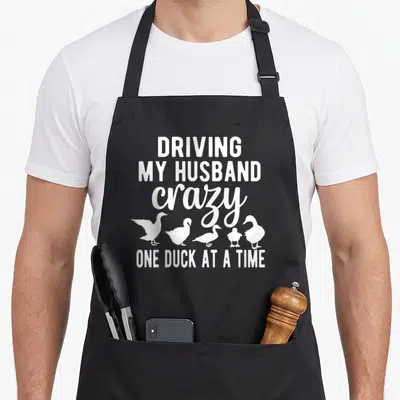 Driving cotton duck aprons - White stacked text Driving My Husband Crazy One Duck At A Time above five duck silhouettes — shown on a black cotton canvas apron against a white background