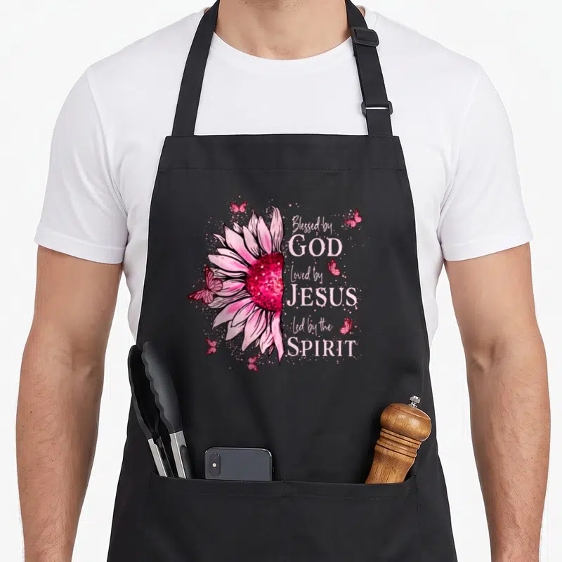 God sunflower apron - Pink watercolor sunflower with butterflies and stacked Christian faith text — shown on a black cotton canvas apron against a white background