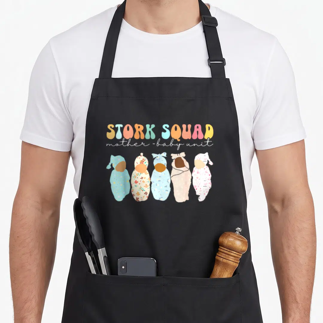Stork my mother's apron - Five swaddled babies in floral and star patterns below STORK SQUAD text in bubble letters — shown on a black cotton canvas apron against a white background