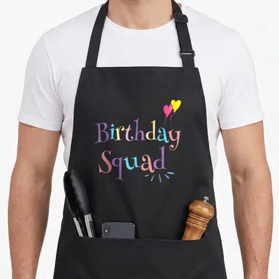Squad pastel birthday apron - Two heart balloons in pink and yellow above multicolored 'Birthday Squad' text — shown on a black cotton canvas apron against a white background
