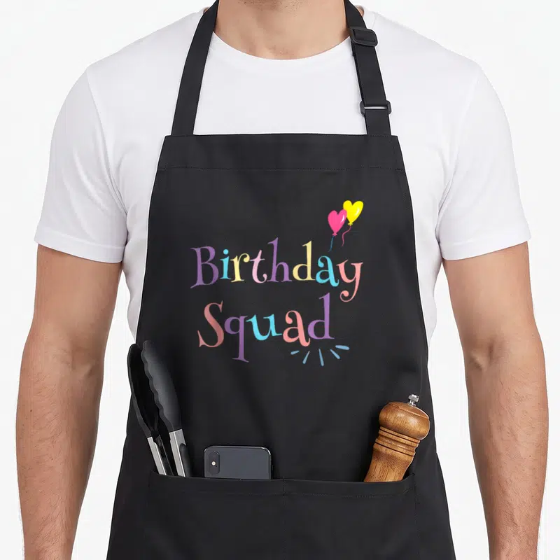 Squad pastel birthday apron - Two heart balloons in pink and yellow above multicolored 'Birthday Squad' text — shown on a black cotton canvas apron against a white background