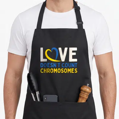 Count chromosomes apron - Love doesn't count chromosomes text with split blue and yellow heart on black background — shown on a black cotton canvas apron against a white background