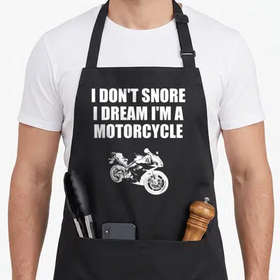 Dream motorcycle apron - White text I DON'T SNORE above a white cartoon sport motorcycle silhouette — shown on a black cotton canvas apron against a white background