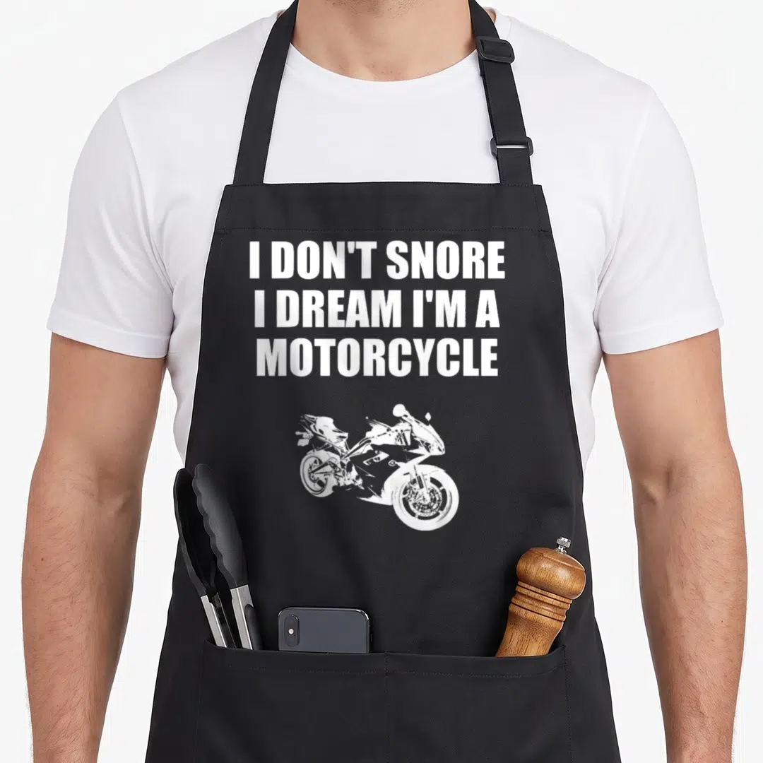 Dream motorcycle apron - White text I DON'T SNORE above a white cartoon sport motorcycle silhouette — shown on a black cotton canvas apron against a white background