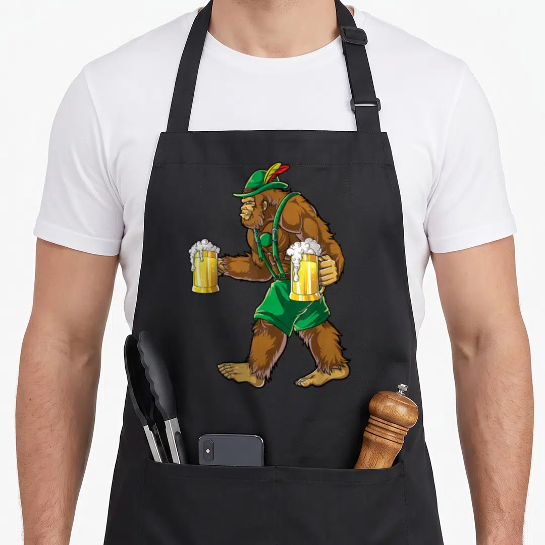 Bigfoot oktoberfest lederhosen apron - Cartoon bigfoot walking in green lederhosen holding two overflowing beer mugs — shown on a black cotton canvas apron against a white background