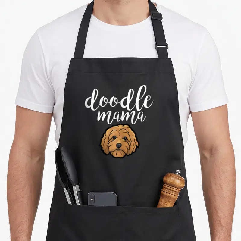 Funny doodle goldendoodle apron - White doodle mama script above a brown cartoon dog head illustration — shown on a black cotton canvas apron against a white background