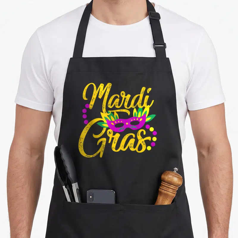 Parade fat chef apron - Yellow Mardi Gras script around a purple masquerade mask with feathers and scattered dots — shown on a black cotton canvas apron against a white background