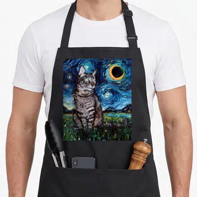 Tabby cat apron - Grey tabby cat sitting in green grass before a swirling blue starry night sky with a large moon — shown on a black cotton canvas apron against a white background
