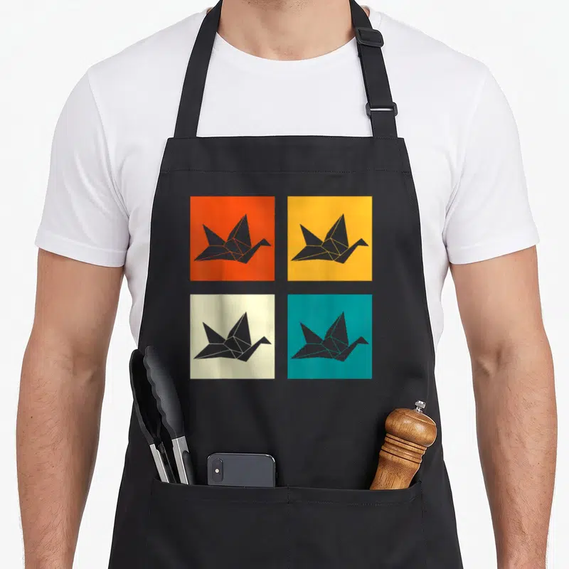 Artist paper apron - Four colored squares with grey origami cranes in orange, yellow, cream, and teal — shown on a black cotton canvas apron against a white background