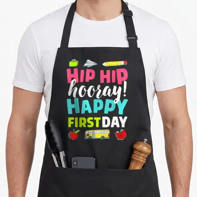 Retro school teacher apron - Cartoon school items and stacked text reading HIP HIP hooray HAPPY FIRST DAY in bright — shown on a black cotton canvas apron against a white background