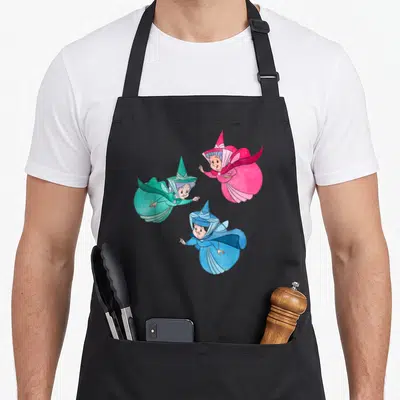 Fairy beauty apron - Three cartoon fairies in pink, green, and blue robes holding wands — shown on a black cotton canvas apron against a white background