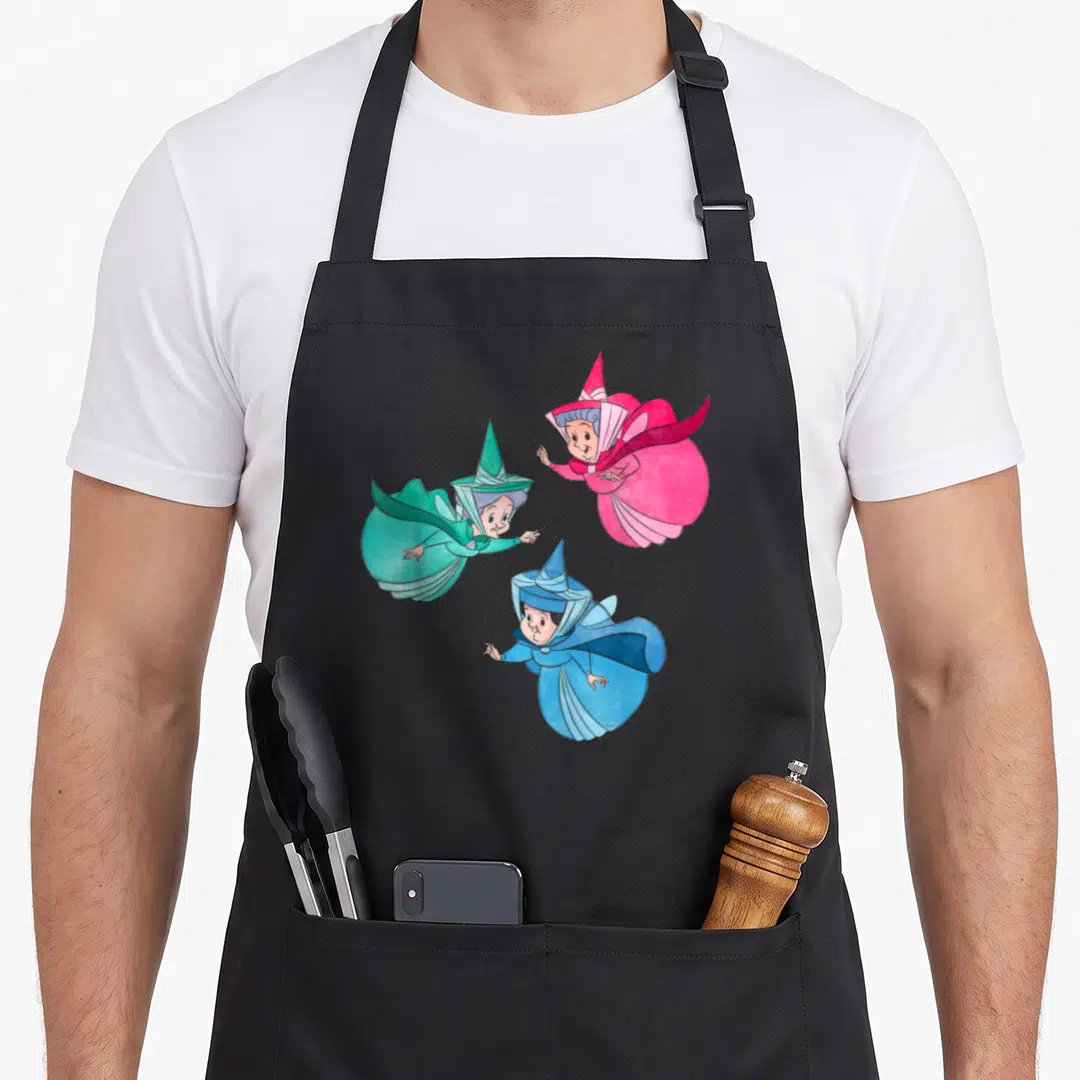 Fairy beauty apron - Three cartoon fairies in pink, green, and blue robes holding wands — shown on a black cotton canvas apron against a white background