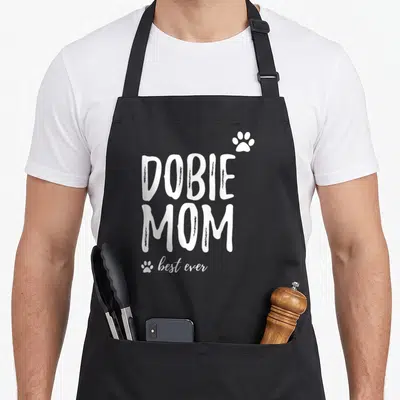 Funny dobie dog apron - White DOBIE MOM text with two paw prints on black bib apron — shown on a black cotton canvas apron against a white background