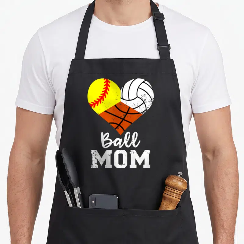 Ball game day apron - Heart made of yellow softball, white volleyball, orange basketball with Ball MOM text — shown on a black cotton canvas apron against a white background