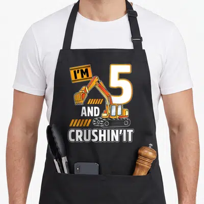 Construction excavator vehicle apron - Cartoon yellow excavator with I'M 5 AND CRUSHIN' IT text and orange stripes — shown on a black cotton canvas apron against a white background