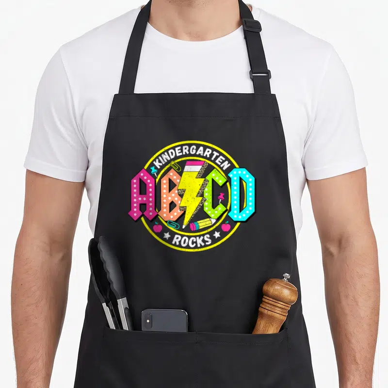 Abcd kindergarten teacher apron - Colorful ABCD letters with pencils and rocks text in yellow circle — shown on a black cotton canvas apron against a white background