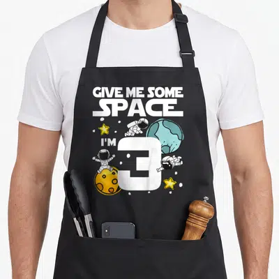Space planet birthday apron - Cartoon astronauts floating near a large white number 3, blue planet, and yellow moon — shown on a black cotton canvas apron against a white background