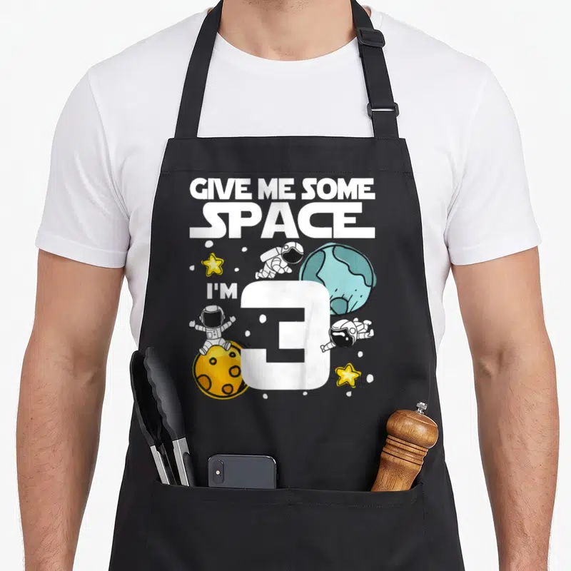 Space planet birthday apron - Cartoon astronauts floating near a large white number 3, blue planet, and yellow moon — shown on a black cotton canvas apron against a white background