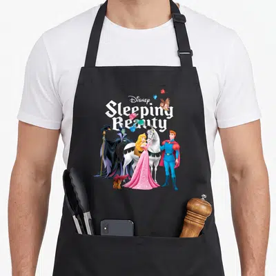 Character beauty apron - Sleeping Beauty title with Aurora, Prince Phillip, and villain in white pink blue purple black — shown on a black cotton canvas apron against a white background