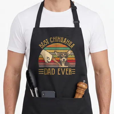 Chihuahua sunset apron - Retro sun with human fist bumping chihuahua paw and arched text reading Best Chihuahua Dad Ever — shown on a black cotton canvas apron against a white background
