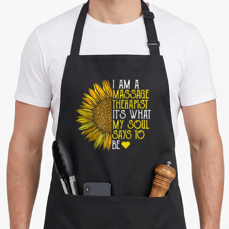 Uniform massage therapist apron - Half sunflower in yellow and brown next to stacked text reading I am a massage — shown on a black cotton canvas apron against a white background