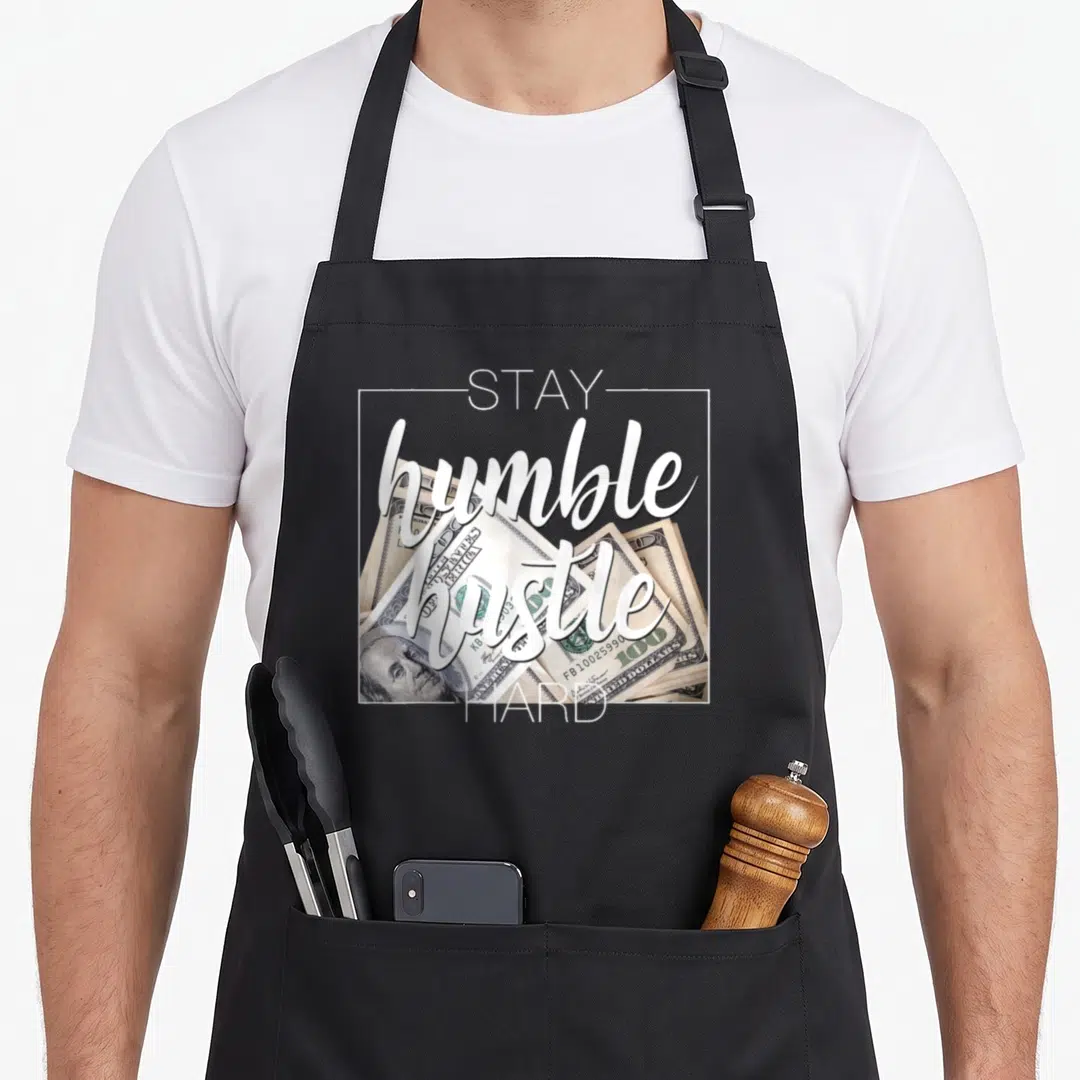 Stay hip apron - Stay Humble Hustle Hard text over stack of 100 dollar bills in white green beige — shown on a black cotton canvas apron against a white background