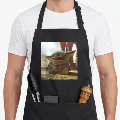 Sitting may look apron - Green character sitting in a robe against a blurred background — shown on a black cotton canvas apron against a white background