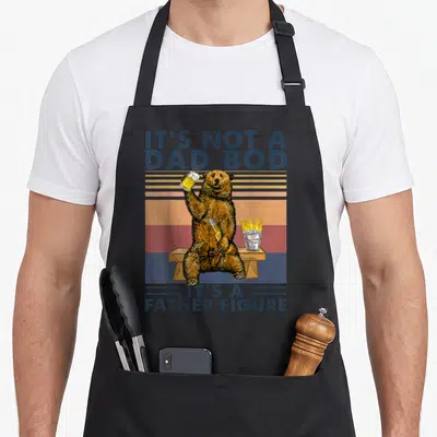 Bod bear apron - Retro bear on bench holding beer glass with ice bucket and father figure text — shown on a black cotton canvas apron against a white background