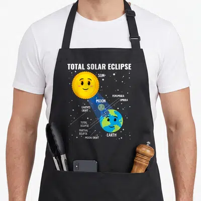 Infographic solar eclipse apron - Cartoon sun and earth with dotted lines labeling penumbra umbra and orbits — shown on a black cotton canvas apron against a white background