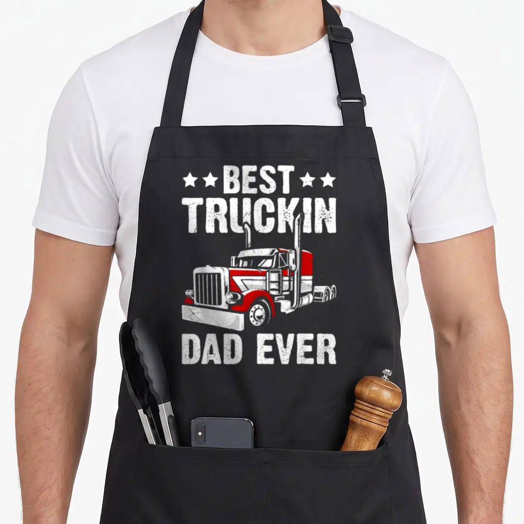 Truckin rig father day apron - Distressed white text BEST TRUCKIN DAD EVER with red and white semi-truck graphic — shown on a black cotton canvas apron against a white background