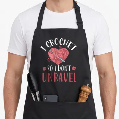 Unravel crochet apron - I CROCHET SO I DON'T UNRAVEL text with red yarn heart and blue hook — shown on a black cotton canvas apron against a white background