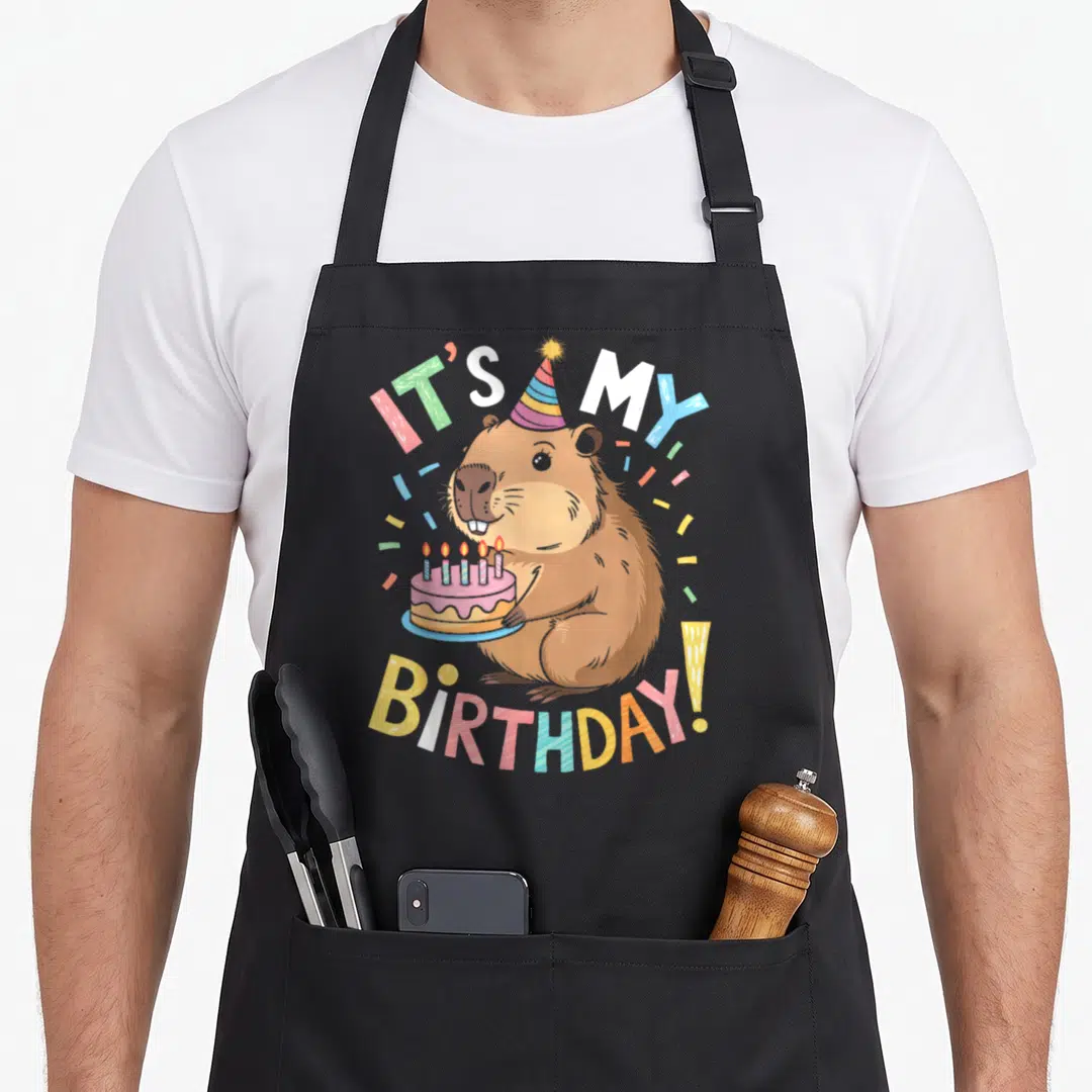 Capybara bday birthday apron - Cartoon brown capybara holding pink cake with lit candles under arching IT'S MY BIRTHDAY — shown on a black cotton canvas apron against a white background