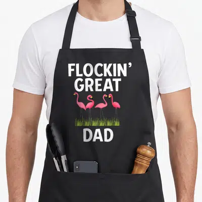 Flockin father day apron - FLOCKIN GREAT DAD text above four pink flamingos standing in green grass — shown on a black cotton canvas apron against a white background