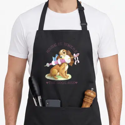 Cute dog lady and the tramp apron - Brown cartoon dog holding pink umbrella with HOME IS WHERE THE DOGS ARE text — shown on a black cotton canvas apron against a white background