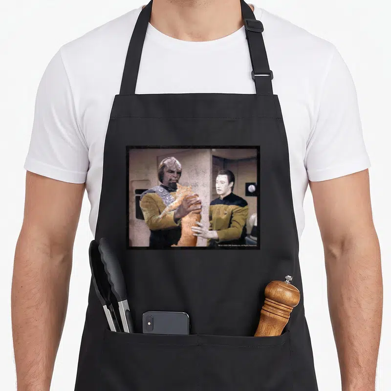 Worf cat apron - Alien holding orange cat next to standing man in photo frame — shown on a black cotton canvas apron against a white background