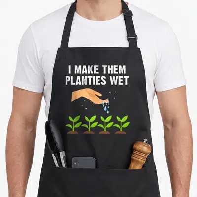 Them plant apron - Cartoon hand dripping blue water onto four green plants in brown soil under white text — shown on a black cotton canvas apron against a white background