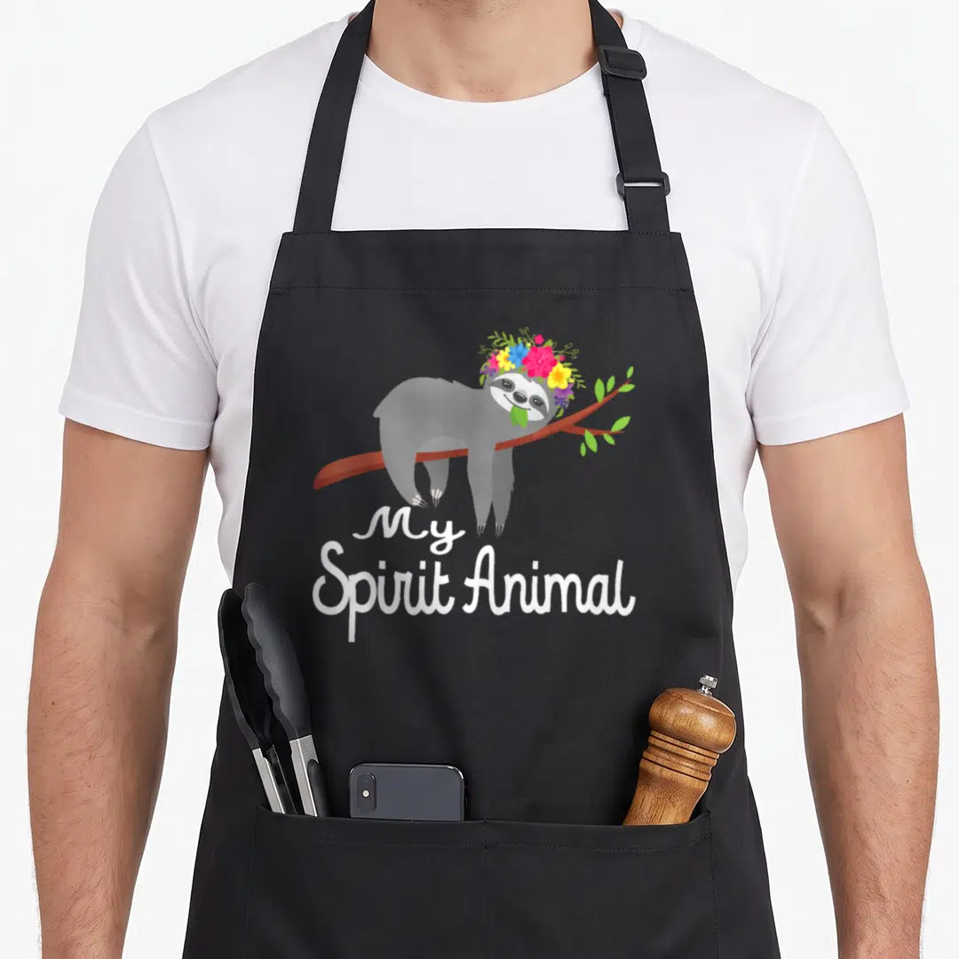 Funny sloth animal apron - Sloth with flower crown hanging below My Spirit Animal text — shown on a black cotton canvas apron against a white background