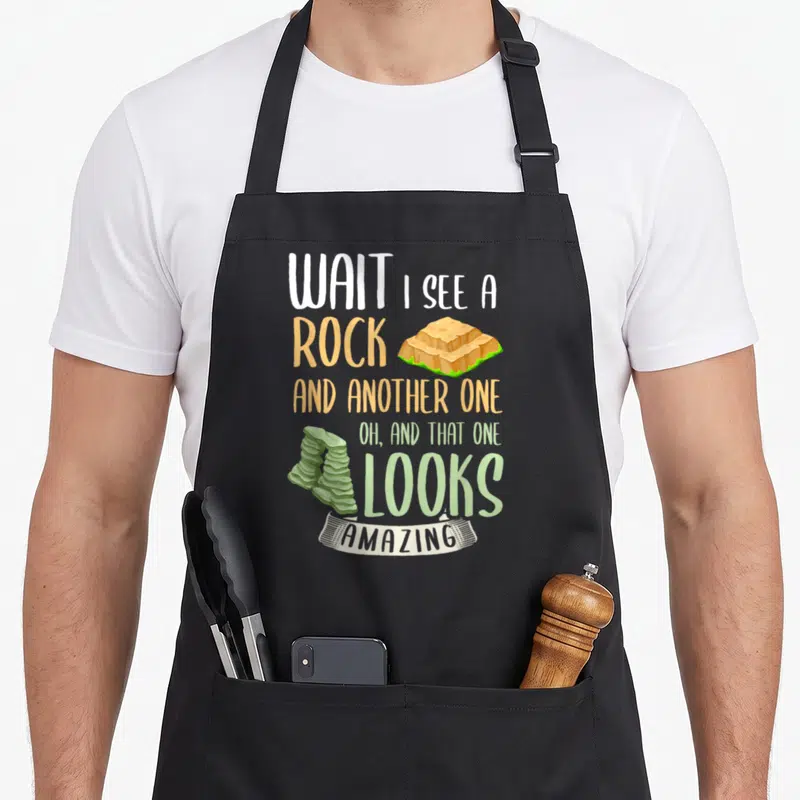 Geologist joke apron - Stacked text reading WAIT I SEE A ROCK with sandwich and sushi stack illustrations in orange — shown on a black cotton canvas apron against a white background