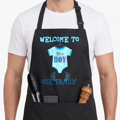 Funny baby shower apron - Blue onesie graphic with It's a BOY text and two footprints — shown on a black cotton canvas apron against a white background