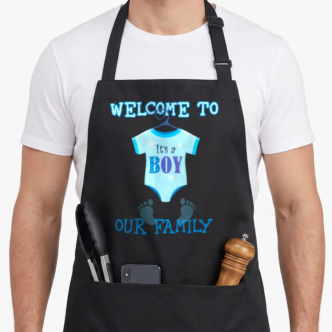 Funny baby shower apron - Blue onesie graphic with It's a BOY text and two footprints — shown on a black cotton canvas apron against a white background