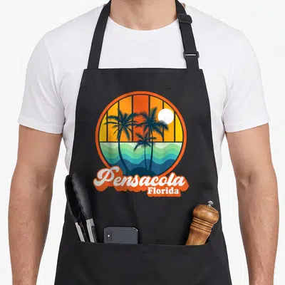 Souvenir summer aprons - Retro circular sunset with palm trees and Pensacola Florida text in orange and blue — shown on a black cotton canvas apron against a white background