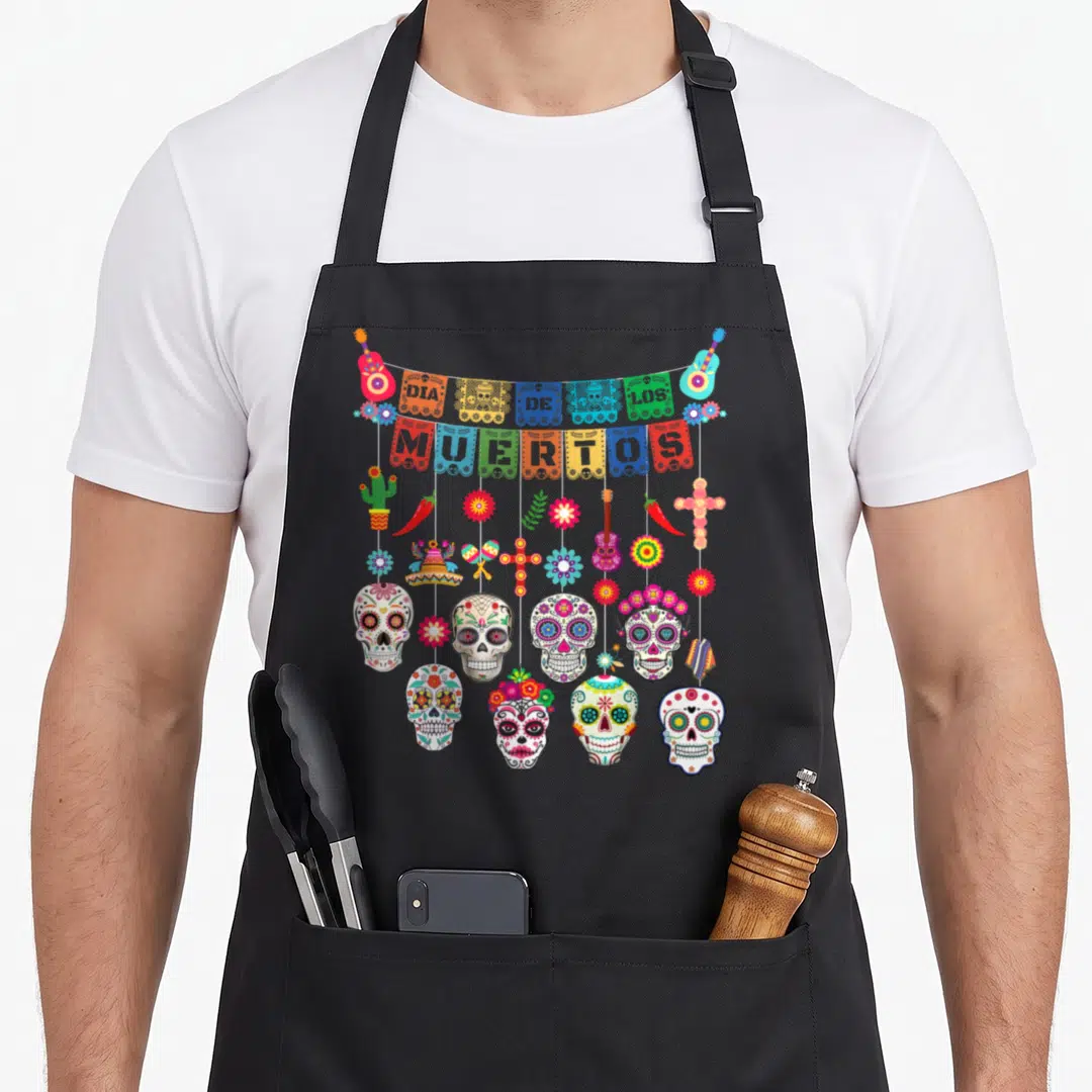 Dia day of the dead apron - Papel picado banners reading DIA DE LOS MUERTOS above hanging maracas, guitars, peppers — shown on a black cotton canvas apron against a white background