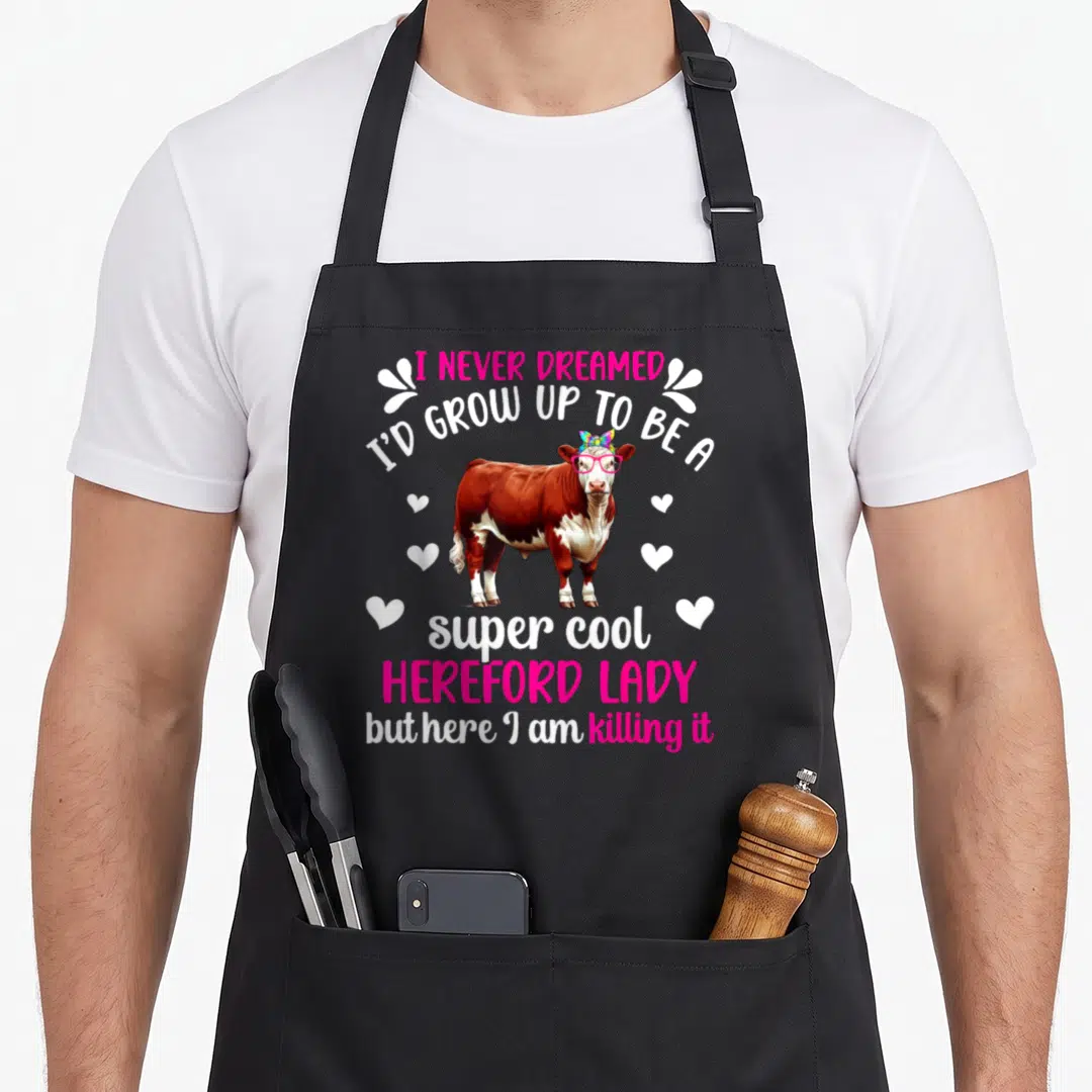 Hereford cow print aprons - Cartoon Hereford cow with pink glasses, rainbow bow, and stacked text surrounded by white — shown on a black cotton canvas apron against a white background