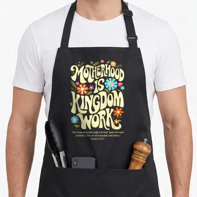 Floral motherhood mother's day aprons - Retro floral typography reading Motherhood is Kingdom Work with Proverbs 31 — shown on a black cotton canvas apron against a white background