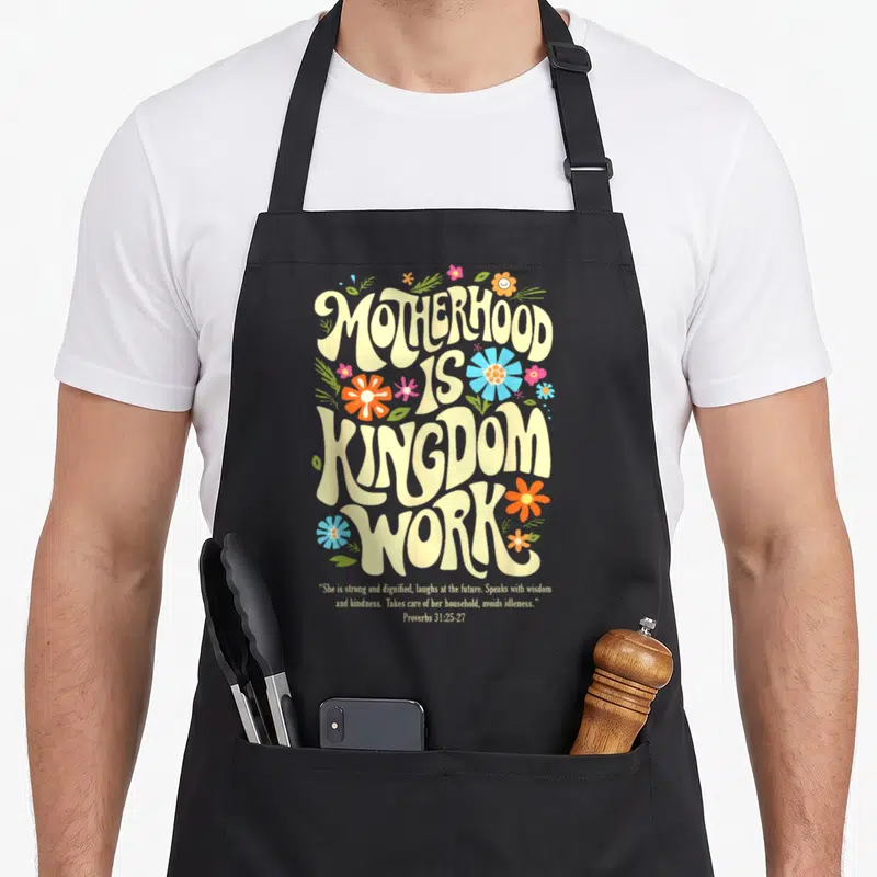 Floral motherhood mother's day aprons - Retro floral typography reading Motherhood is Kingdom Work with Proverbs 31 — shown on a black cotton canvas apron against a white background