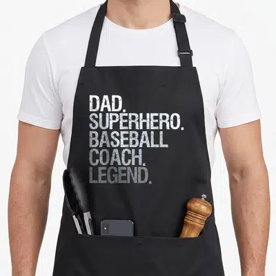Funny coach baseball apron - White stacked text reading dad superhero baseball coach legend on black background — shown on a black cotton canvas apron against a white background