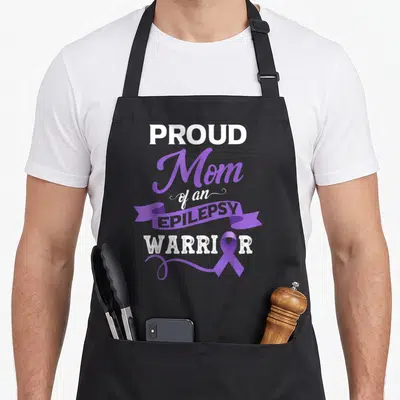Month epilepsy awareness apron - Stacked text proud mom of an epilepsy warrior with purple ribbon and banner — shown on a black cotton canvas apron against a white background
