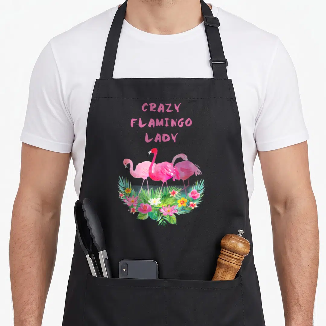 Collector pink flamingo apron - Three pink flamingos behind tropical flowers with stacked pink text reading CRAZY — shown on a black cotton canvas apron against a white background