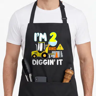 2nd birthday truck apron - Yellow front loader with I'M 2 AND DIGGIN' IT text and construction safety gear — shown on a black cotton canvas apron against a white background