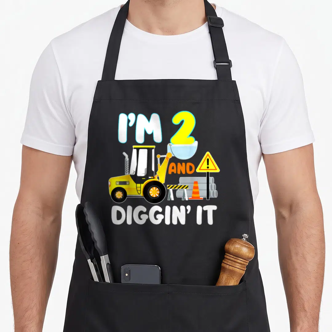2nd birthday truck apron - Yellow front loader with I'M 2 AND DIGGIN' IT text and construction safety gear — shown on a black cotton canvas apron against a white background